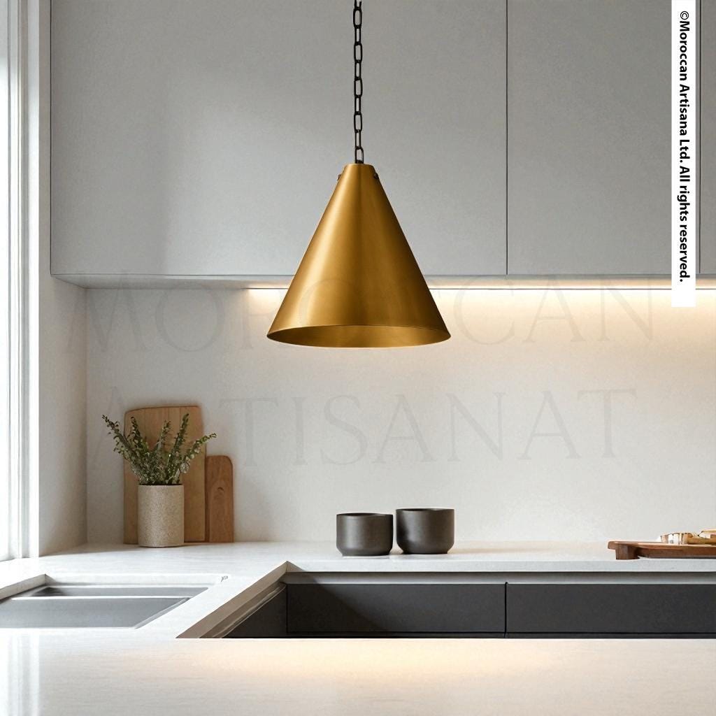 a modern kitchen with a sleek, minimalist design. The focal point is a gold pendant light hanging above a white countertop, which is adorned with a potted plant and a wooden cutting board. The kitchen also includes a sink and a set of black bowls on the countertop.