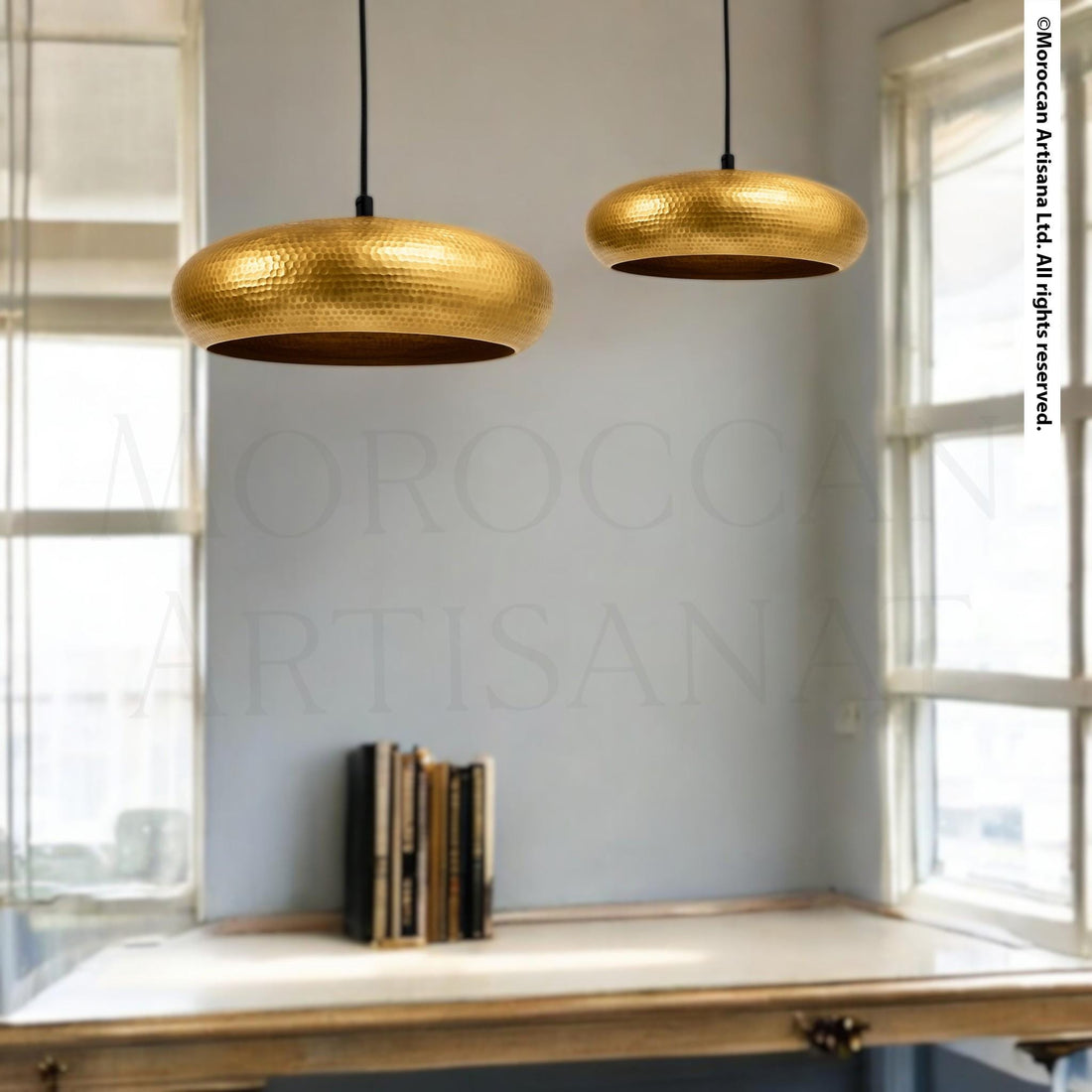 two gold-colored pendant lights hanging from the ceiling, illuminating a room with a white wall and a window. In the background, there is a wooden desk with a stack of books on it.