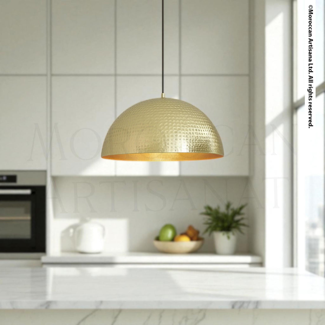 a modern kitchen with a large, gold-colored pendant light hanging above a marble countertop. The countertop has various kitchen items, including a bowl of fruit, a teapot, and a potted plant. The overall design and color scheme of the kitchen create a stylish and contemporary atmosphere.