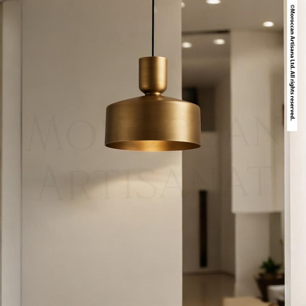 A modern gold-colored pendant light hangs from the ceiling in a minimalist room with white walls and a wooden floor.