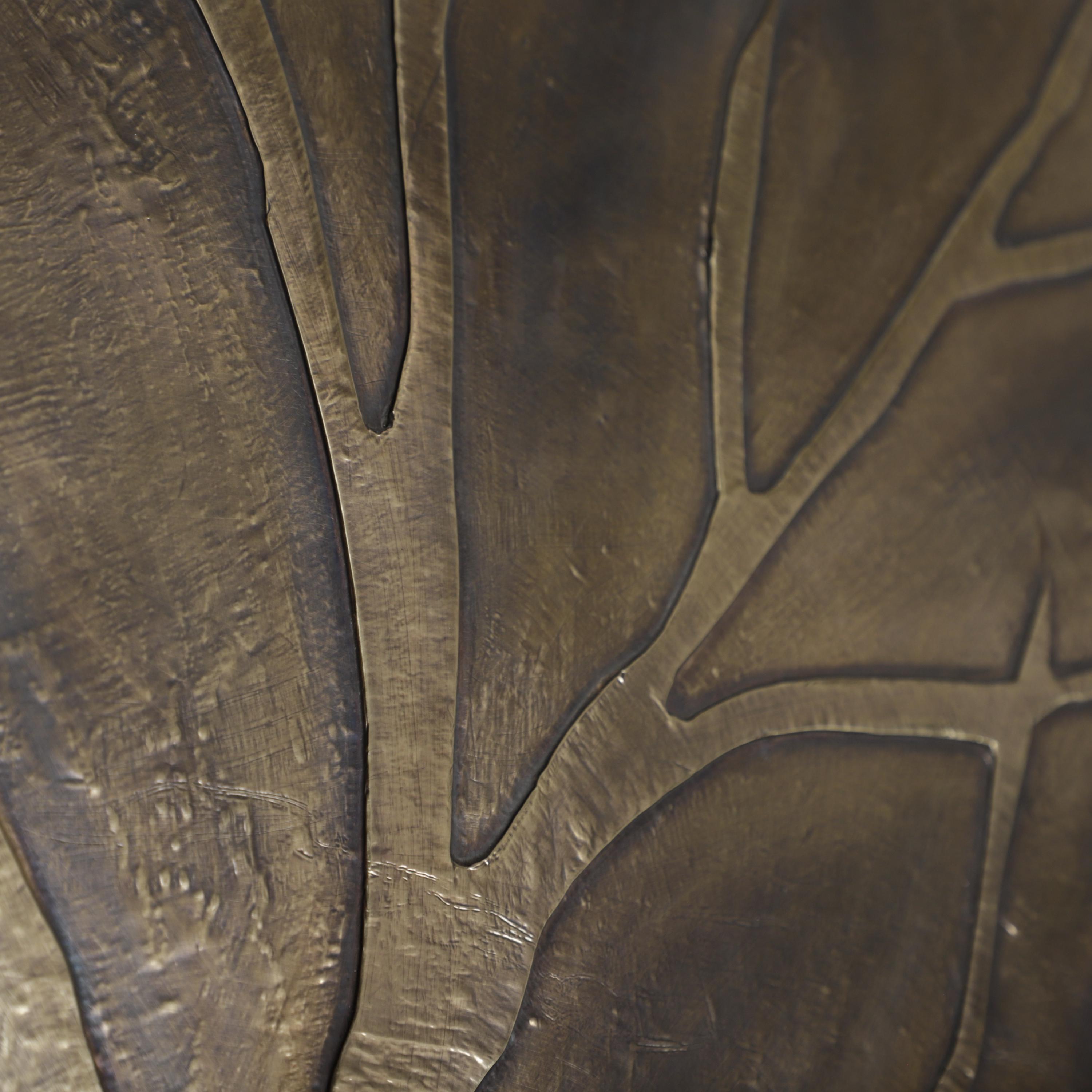 a close-up view of a textured surface, possibly a piece of metal or a fabric, with a pattern of lines and shapes. The surface appears to be dark in color, possibly bronze or a similar shade. The image does not contain any text or other discernible objects or actions.