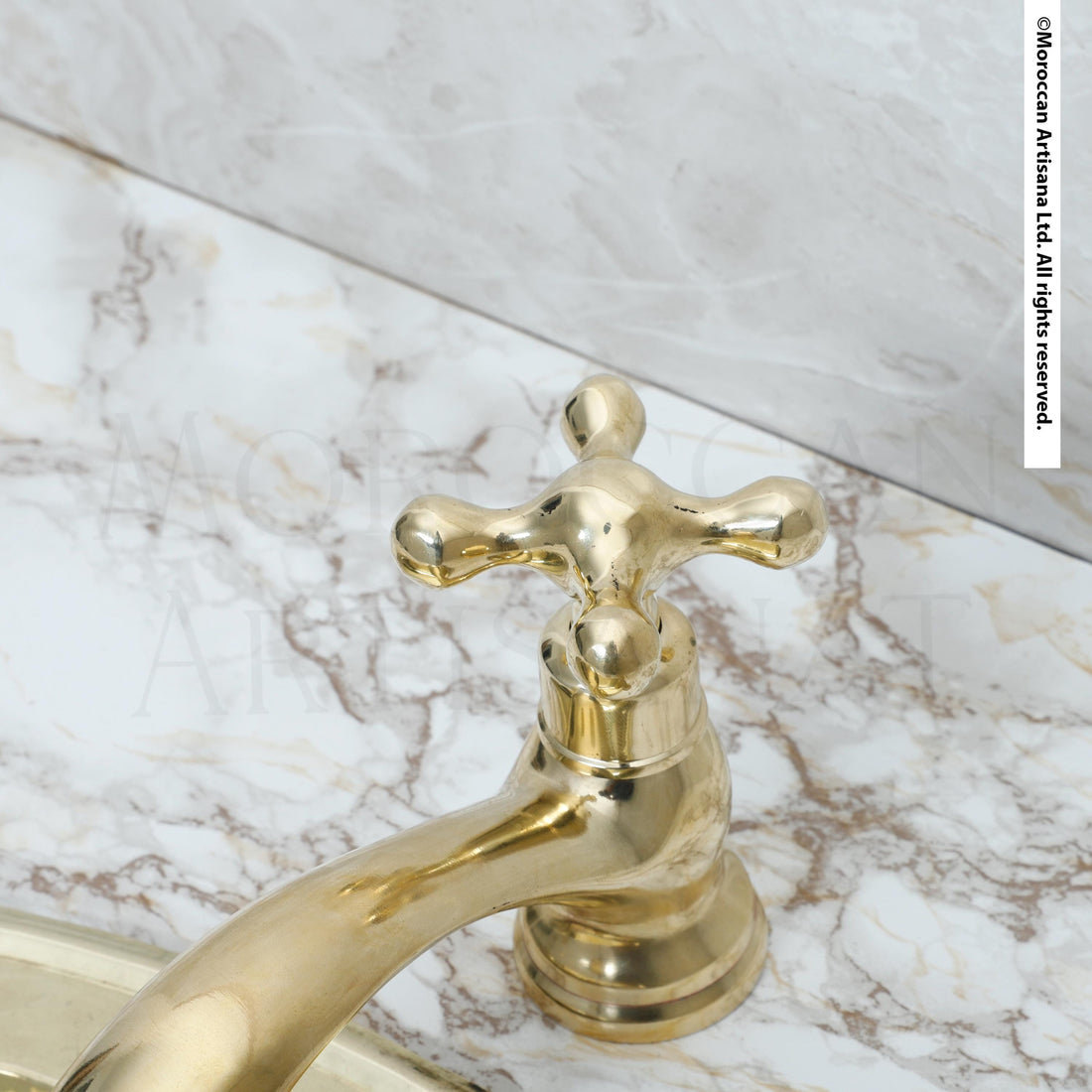 A close-up view of a gold-colored bathroom faucet with a cross-shaped handle, set against a marble countertop.