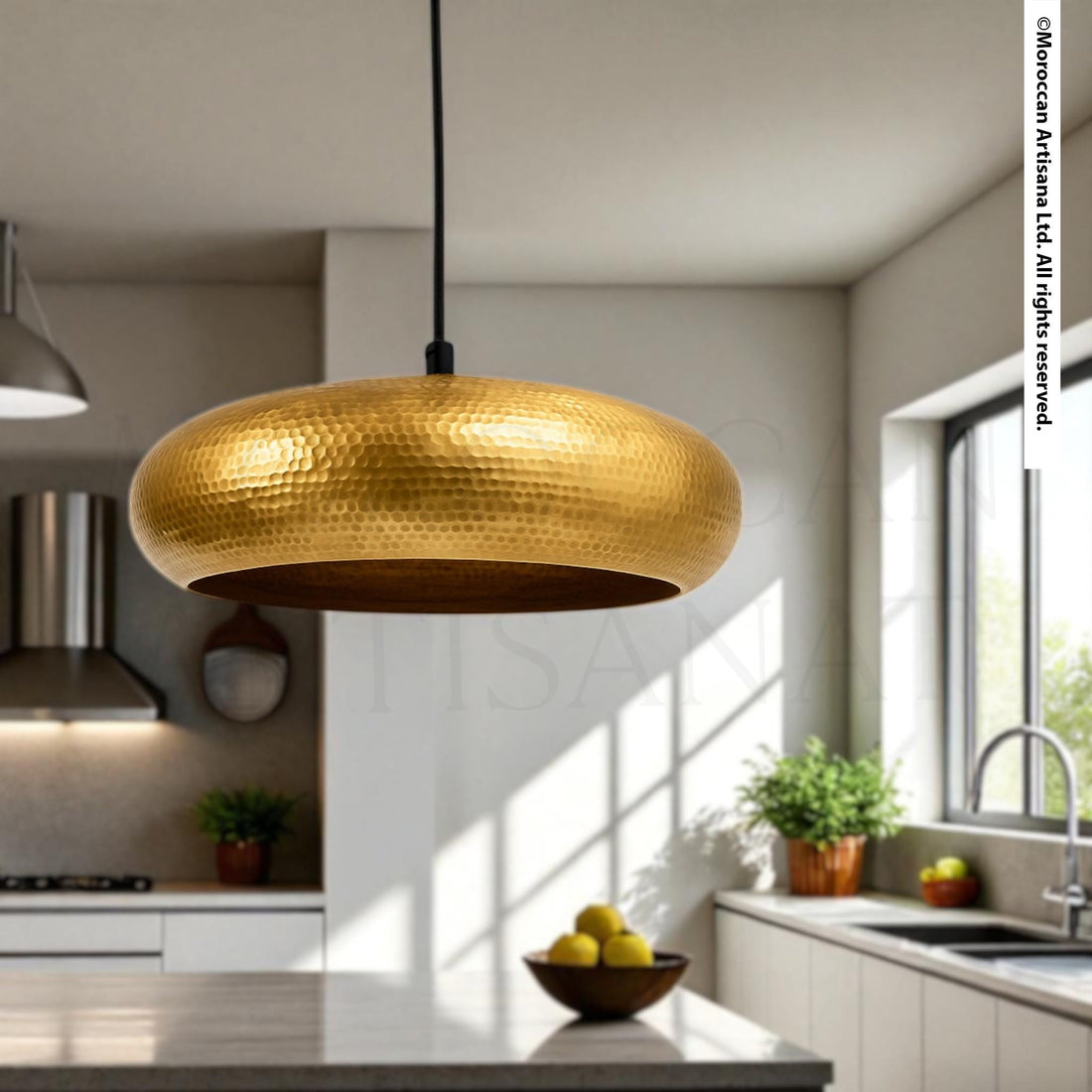 a modern kitchen with a large, gold-colored pendant light hanging from the ceiling, casting a warm glow over the space. The kitchen is well-lit, with a sink and various decorative elements such as potted plants and a bowl of fruit on the counter.