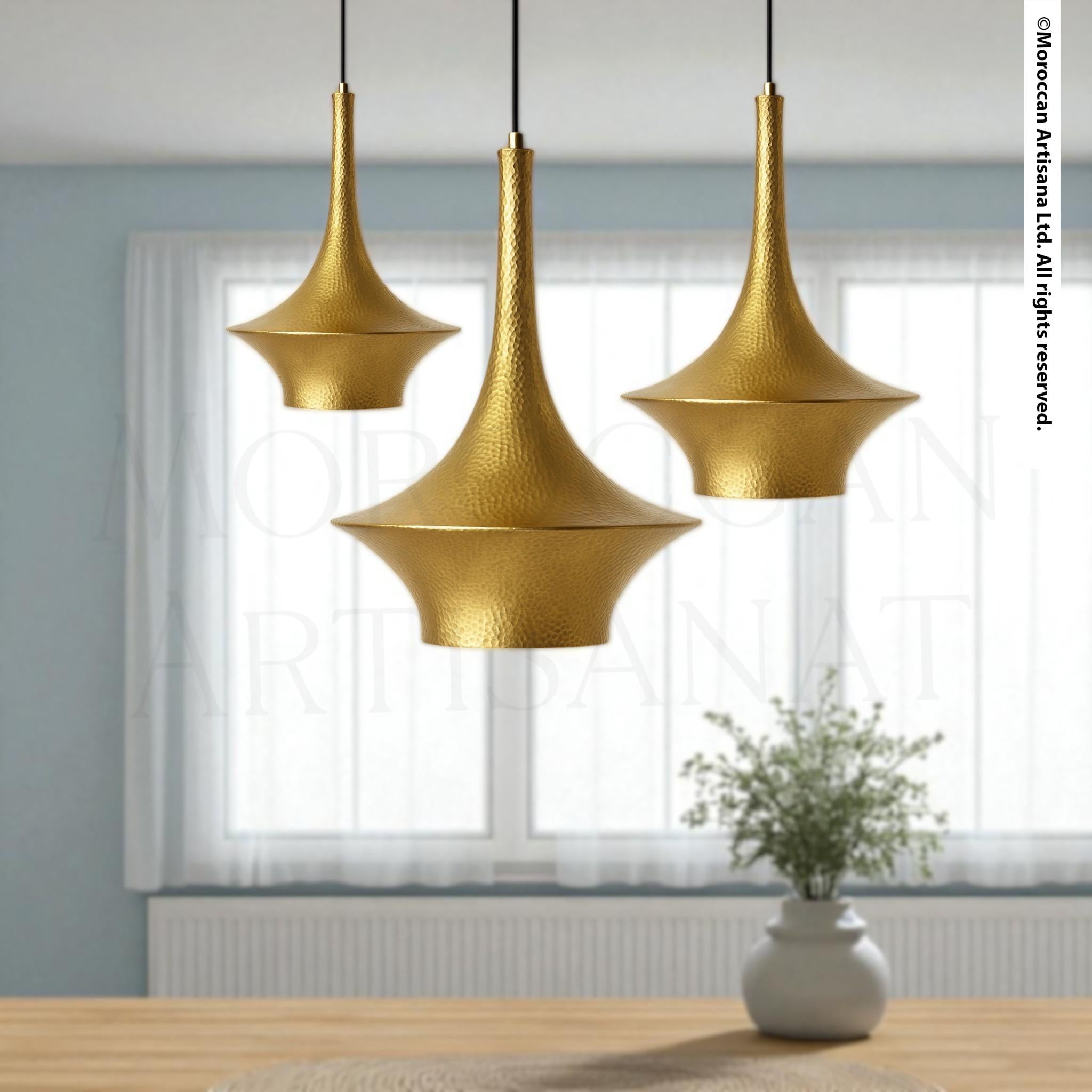 a modern, minimalist interior design with three gold-colored pendant lights hanging from the ceiling, casting a warm glow over the room. In the background, there is a window with white curtains, and a potted plant placed on a wooden table, adding a touch of greenery to the space.