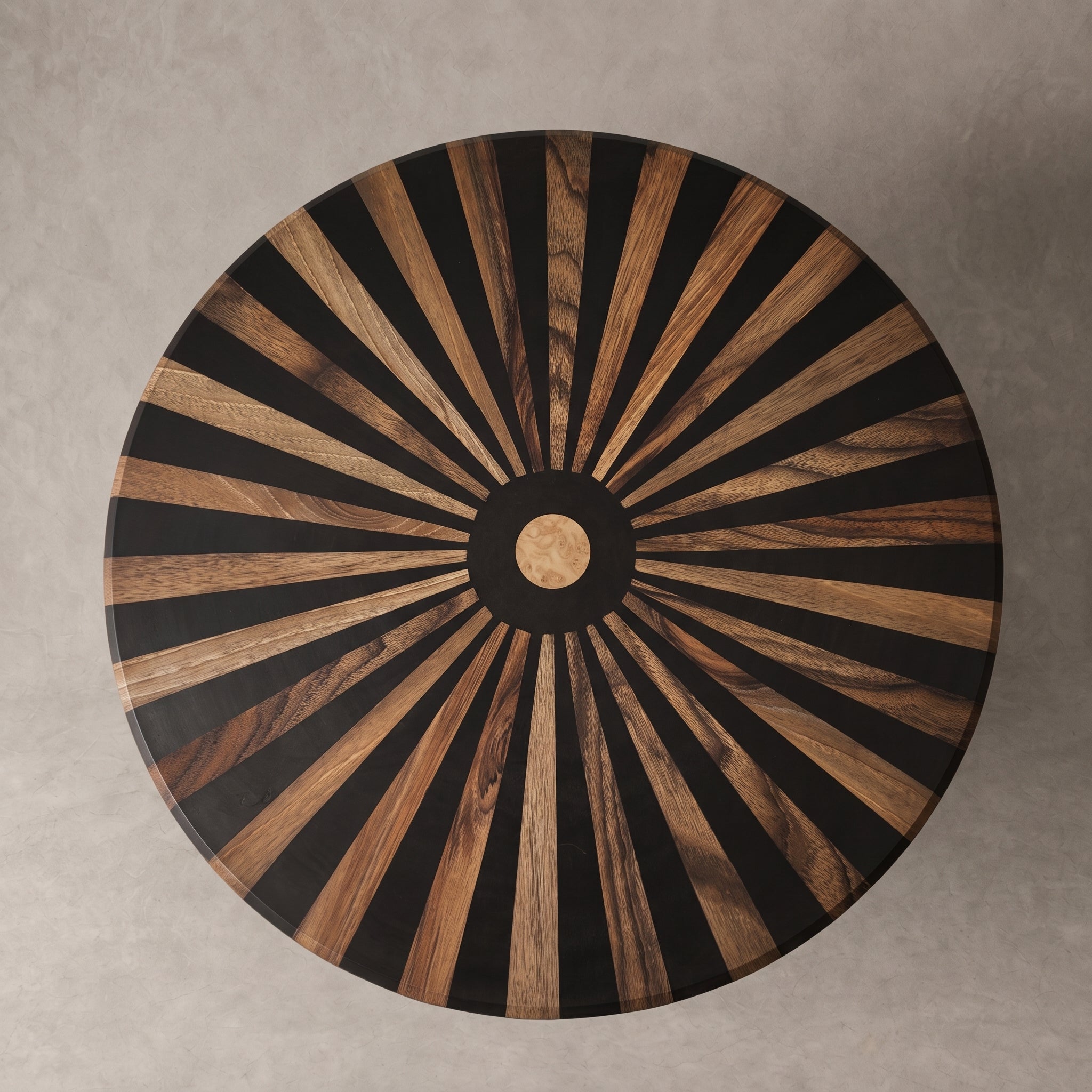 A round wooden table top viewed from directly above, featuring a radial sunburst inlay pattern of alternating black and warm brown wood rays, with a circular wood center medallion, set against a neutral gray concrete background.