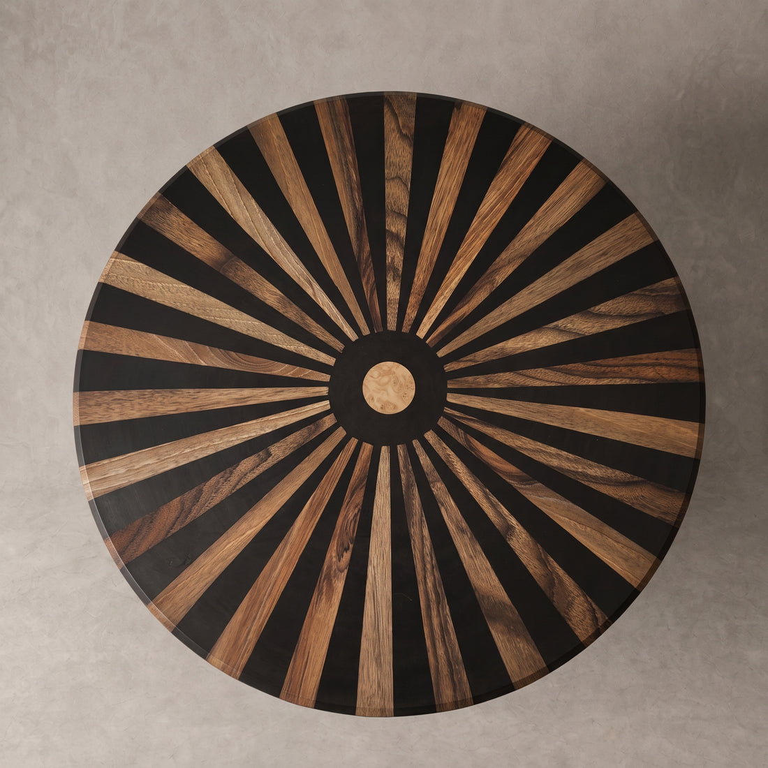 A round wooden table top viewed from directly above, featuring a radial sunburst inlay pattern of alternating black and warm brown wood rays, with a circular wood center medallion, set against a neutral gray concrete background.