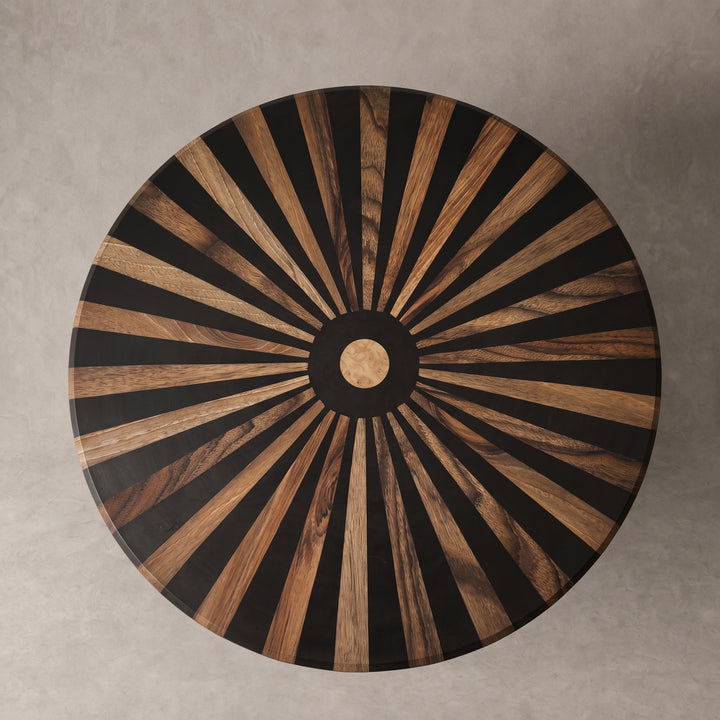 A round wooden table top viewed from directly above, featuring a radial sunburst inlay pattern of alternating black and warm brown wood rays, with a circular wood center medallion, set against a neutral gray concrete background.
