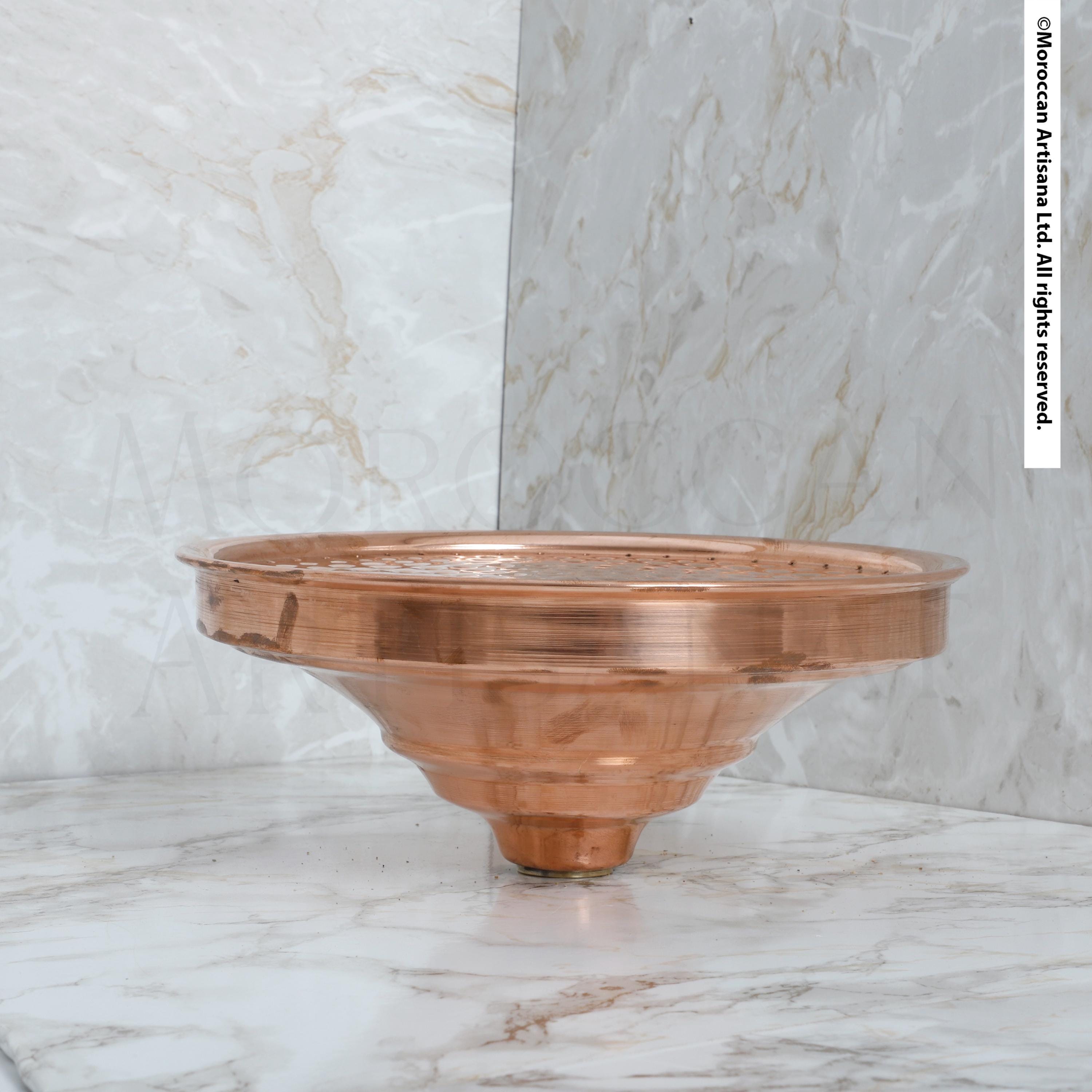 A copper-colored metal object, possibly a bowl or vase, is placed on a marble surface against a marble wall.