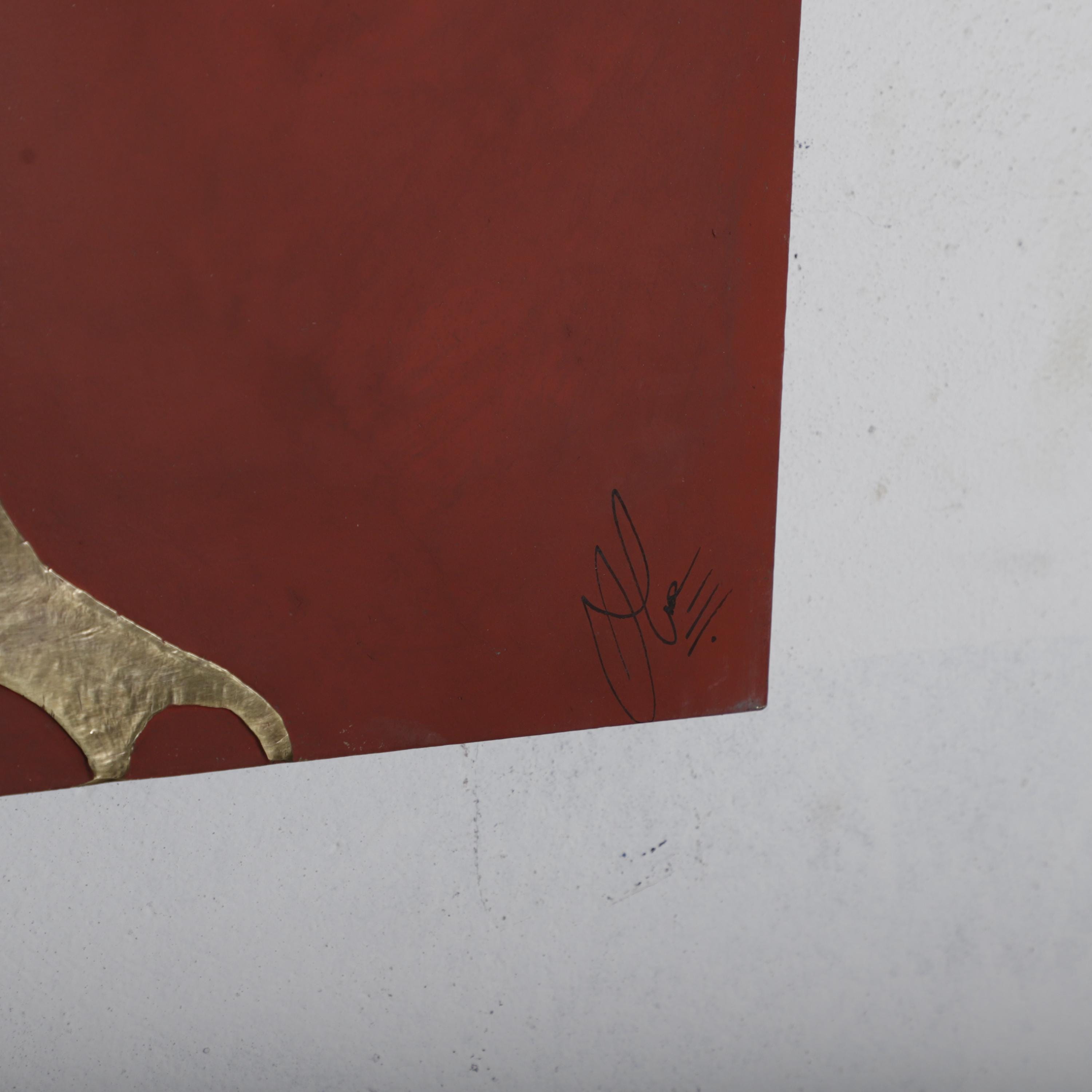 The image is a close-up of a red surface with a gold-colored object, possibly a sculpture or artwork. There is a signature or mark in the bottom right corner of the red surface.
