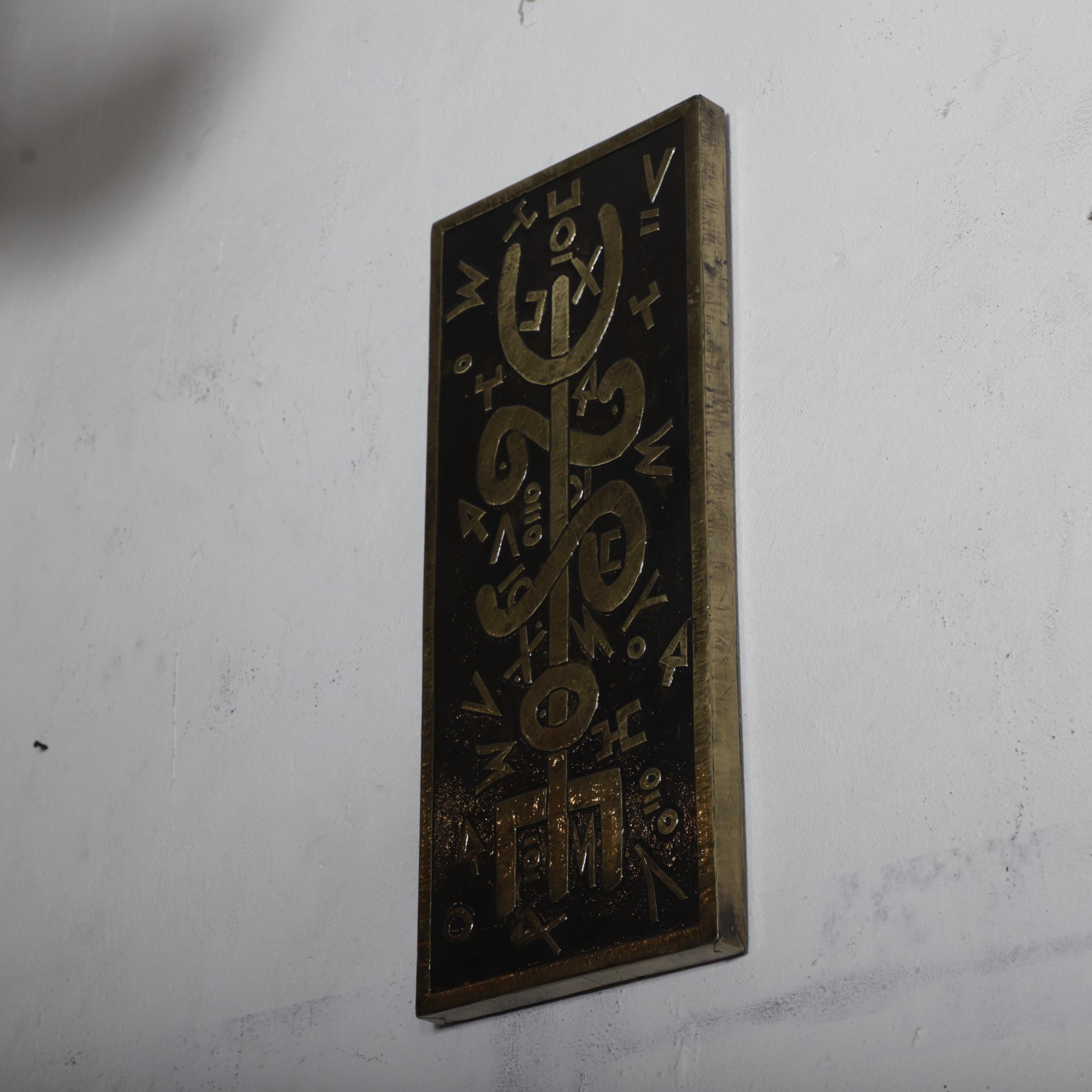 a rectangular metal plaque with an intricate design and text on it, mounted on a white wall.