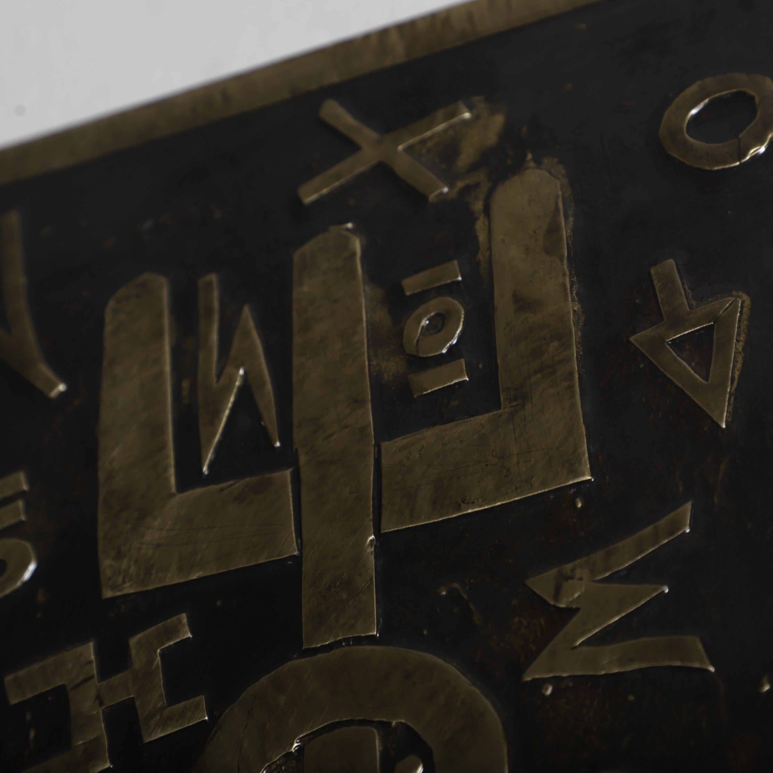 a close-up view of a dark metal surface with various metallic symbols and shapes, including a large &quot;L&quot; and other geometric designs.