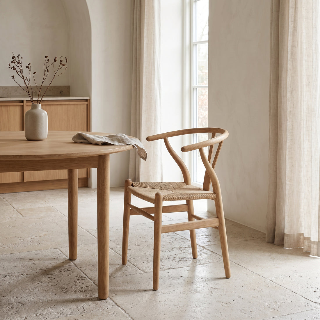 A light natural wood dining table and a matching woven-back chair in a bright, minimalist room with beige walls, stone floors, and sheer curtains.