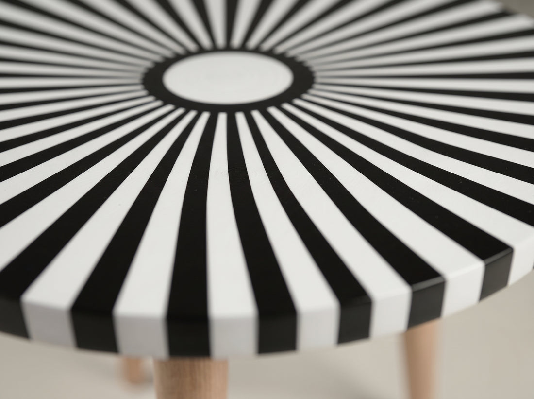 A close-up of a round table with a bold black and white sunburst pattern on the tabletop and light wood legs.