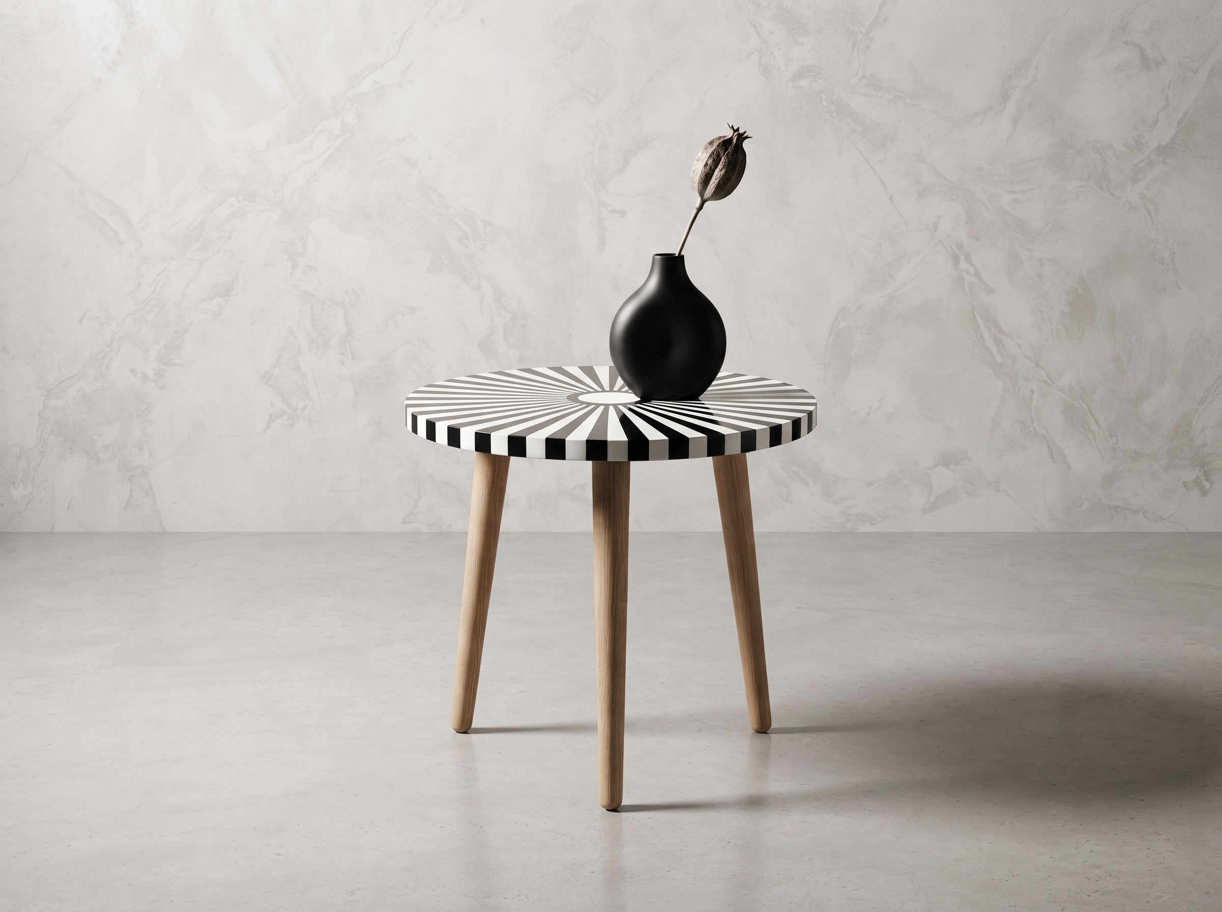 A round side table with a black and white striped top and light wood legs, styled with a black ceramic vase holding a dried flower, set against a gray marble-textured wall and concrete floor.