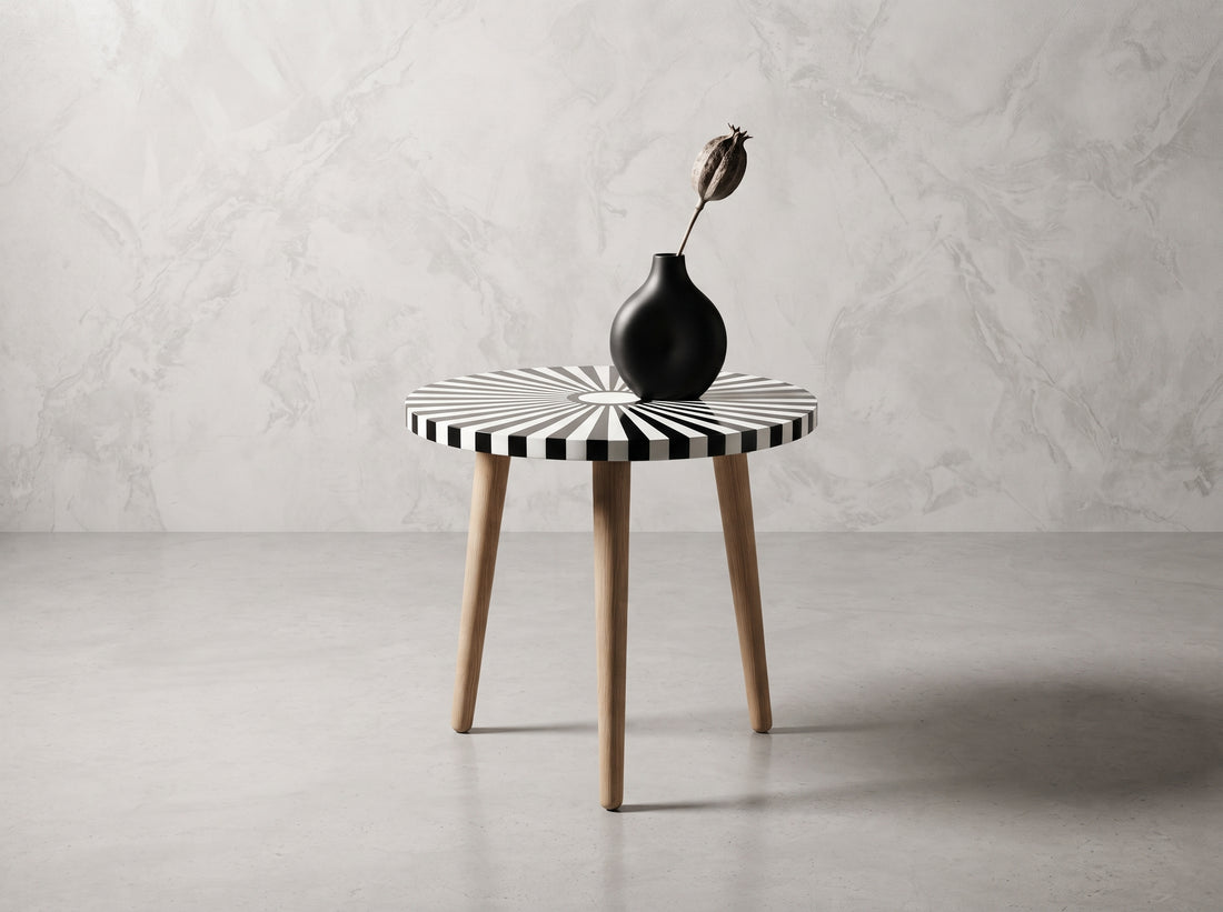 A round side table with a black and white striped top and light wood legs, styled with a black ceramic vase holding a dried flower, set against a gray marble-textured wall and concrete floor.