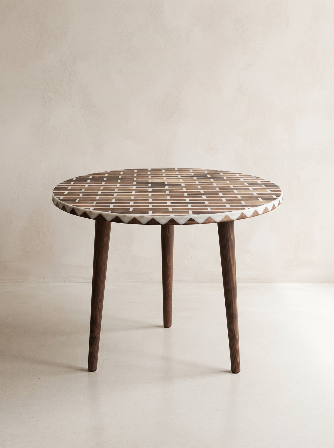 A round wooden dining table with a geometric mosaic inlay top and three tapered dark wood legs, set against a neutral beige textured wall.