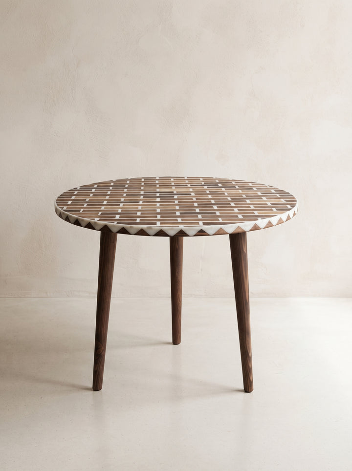A round wooden dining table with a geometric mosaic inlay top and three tapered dark wood legs, set against a neutral beige textured wall.