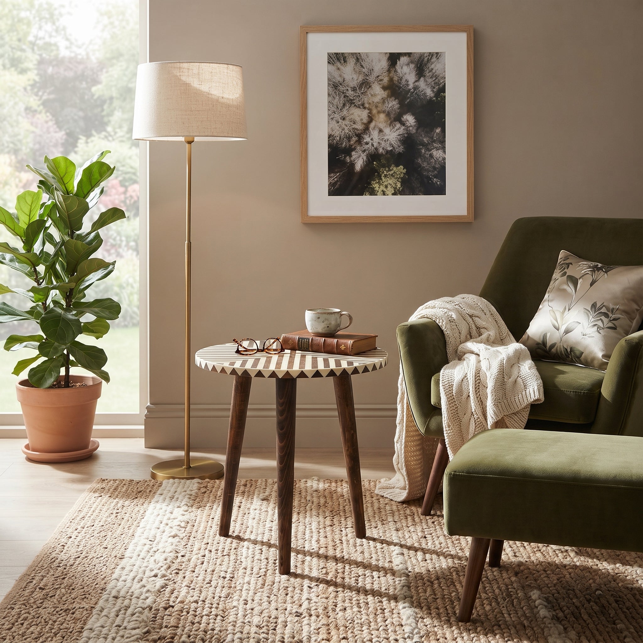 A styled living room corner featuring a round wooden side table with a geometric patterned top, next to an olive green armchair with a cream throw blanket and a matching ottoman. A tall floor lamp stands near a window next to a potted fiddle-leaf fig plant, and framed wall art hangs on the beige wall.