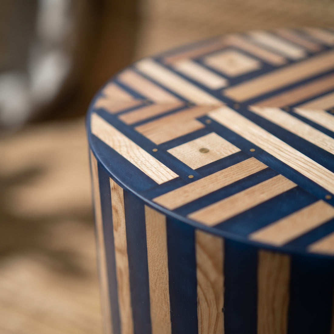 Close-up of a cylindrical wooden stool or side table with a geometric inlay pattern on the top and alternating dark blue and natural wood vertical stripes on the side, photographed in a warm indoor setting.