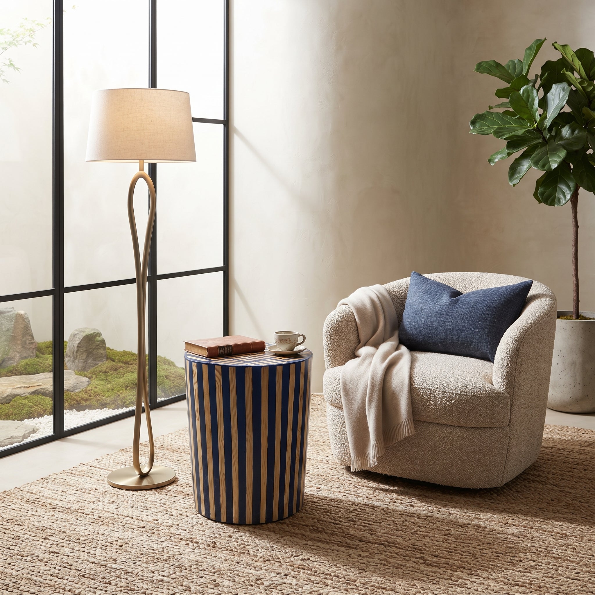 A styled interior scene with a cream boucle armchair, a navy blue accent pillow, a striped side table, a floor lamp, and a potted plant in a sunlit room.