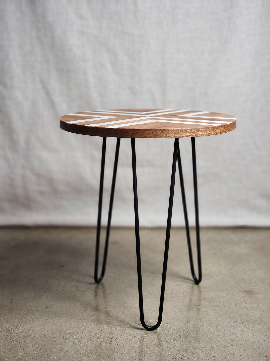 A round wooden side table with a natural wood top featuring a white geometric line pattern, supported by three black metal hairpin legs, set against a neutral gray fabric backdrop.